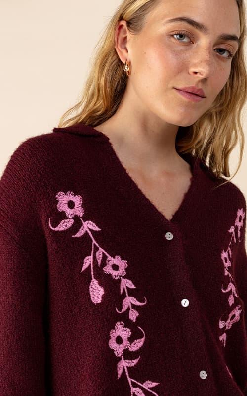 A person wearing a maroon cardigan with pink floral embroidery and pearl buttons, standing against a neutral background.