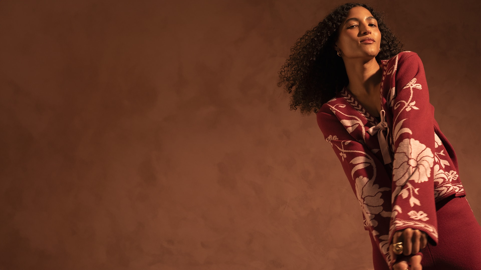 A woman with curly hair wearing a maroon floral-patterned cardigan poses confidently against a brown background.